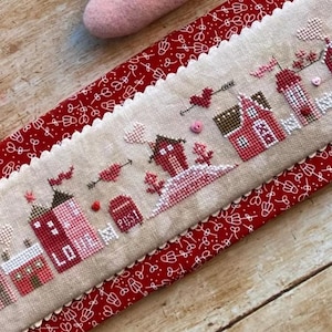 May include: A cross-stitch sampler featuring a row of red and pink houses, trees, and hearts. The word "LOVE" is stitched on one house. The sampler is on a beige background with a scalloped edge, set on a red fabric with a white floral pattern.