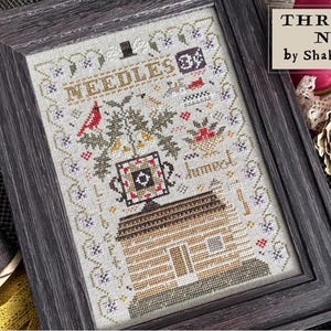 May include: Framed cross-stitch sampler with a rustic cabin design. The artwork features a red cardinal, floral elements, and the text "Needles 3/4" and "Homet". The frame is a dark gray wood.