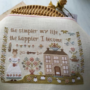 May include: A cross-stitch sampler with a quote that reads "The simpler my life, the happier I become". The sampler features a small house, a woman holding a cup, and a garden with flowers and sheep.