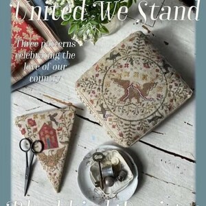 May include: A photograph showcasing cross-stitch patterns with the text "United We Stand" and "Blackbird Designs." The patterns depict a house, birds, and floral motifs in muted tones. Scissors and sewing notions are also present.