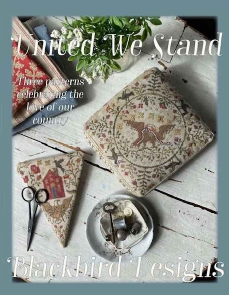 May include: A photograph showcasing cross-stitch patterns with the text "United We Stand" and "Blackbird Designs." The patterns depict a house, birds, and floral motifs in muted tones. Scissors and sewing notions are also present.