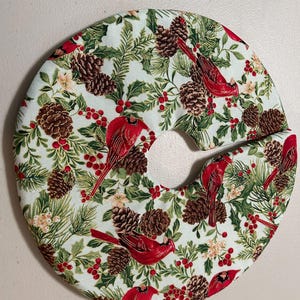 May include: A circular, padded item with a festive Christmas design. The fabric features red cardinals, pine cones, holly, and berries on a light green background. The item has a curved cutout.