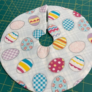 May include: A white fabric Easter tree skirt with a hole in the center for a tree trunk. The skirt is decorated with colorful Easter eggs in various patterns, including polka dots, stripes, and checkered designs.