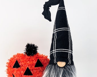 May include: A black and white plaid gnome with a black bat wing hat and a white beard. The gnome is holding a red and black jack-o'-lantern.