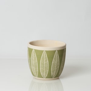 May include: A green ceramic planter with a white leaf pattern. The planter is round and has a wide opening.