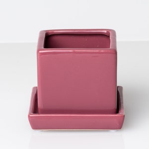 May include: A square, ceramic planter in a muted rose color. The planter has a matching square saucer. The glossy finish reflects light. This decorative planter is suitable for small plants and succulents.