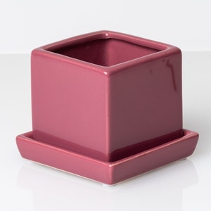 May include: A square, magenta ceramic plant pot with a matching square saucer. The glossy finish reflects light, highlighting the clean lines of the modern design. Suitable for small plants or succulents. The pot is approximately 10 cm square.
