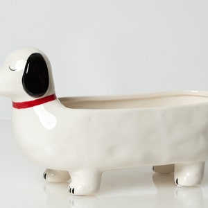 Puede incluir: Maceta de cerámica blanca con forma de dachshund, con orejas negras y un collar rojo. La maceta tiene un cuerpo largo y rectangular y cuatro patas cortas.