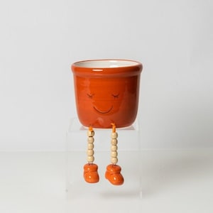 Peut inclure: Pot en céramique orange avec un visage souriant et des pieds en bois. Le pot est posé sur un support en acrylique transparent.