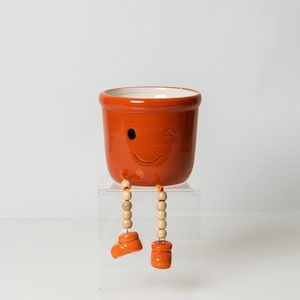 May include: Orange ceramic planter with a winking smiley face and wooden legs. The planter is sitting on a clear stand.