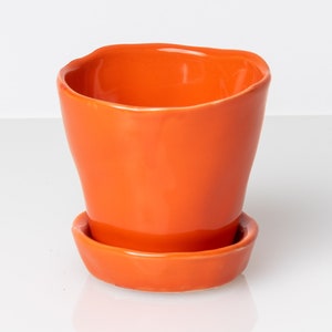 May include: An orange ceramic planter with a matching saucer. The planter has a slightly irregular, curved rim and a smooth, glossy finish. The design is simple and modern, suitable for small plants or succulents.