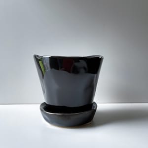 May include: A black ceramic plant pot with a matching saucer. The pot has an unusual, slightly uneven shape and a glossy finish. The saucer is round and sits beneath the pot, creating a cohesive look. Suitable for small plants or succulents.
