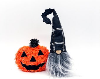 May include: A black and white plaid gnome with a black bat on its hat and a long white and grey beard. The gnome is standing next to a bright orange jack-o'-lantern with a black face.
