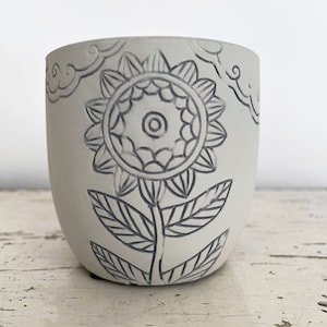 May include: White ceramic planter with a black and white floral design. The planter features a stylized sunflower with leaves.