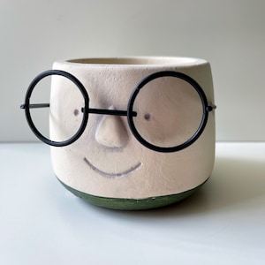 以下が含まれることがあります： 黒い丸い眼鏡と緑色の縁取りが付いた顔の形をした白いセラミック製のプランター