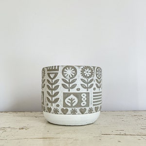 May include: A white ceramic planter with a gray geometric pattern of flowers, leaves, and hearts. The planter has a white base.