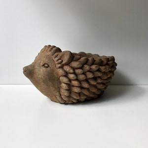 May include: A brown ceramic hedgehog planter with a detailed face and textured body. The planter is designed with a shallow bowl shape, perfect for small plants or succulents. The hedgehog's face has small, detailed eyes and a pointed snout.