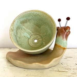 Puede incluir: Un macetero de caracol de cerámica verde glaseada con un borde marrón y un pequeño agujero de drenaje en la parte inferior. El caracol sostiene una maceta de cerámica verde glaseada con un borde marrón.