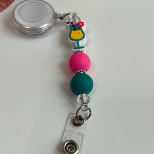 May include: A retractable badge reel with a clear plastic strap and a cocktail-themed charm. The charm features a yellow cocktail in a glass, a pink flower, and colorful beads in pink and teal. The reel is silver.