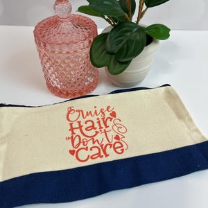 May include: A canvas cosmetic bag with the text "Cruise Hair Don't Care" in red script. The bag has a cream-colored body with a navy blue border. A pink glass jar with a lid and a small potted plant are also in the image.