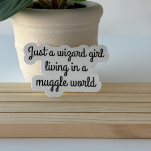 May include: A white die-cut sticker featuring black cursive text that reads, Just a wizard girl living in a muggle world. The sticker has a smooth, rounded border and is displayed against a neutral background, showcasing clear, legible typography.