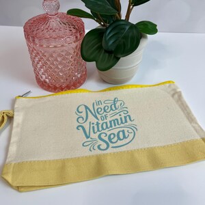 May include: A canvas cosmetic bag with a yellow trim and the text "in Need of Vitamin Sea" in teal script. A pink glass jar with a lid and a small potted plant are also in the image.