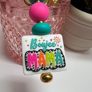 May include: A keychain with a white square pendant that reads "Boujee MAMA" in colorful, bubble letters. The keychain features turquoise and pink beads, a gold-colored chain, and a gold-colored bead at the bottom.
