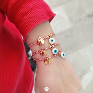 May include: Three red string bracelets with gold charms. The charms are a ladybug, a small square, and a blue evil eye.
