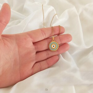Puede incluir: Un collar de cadena dorada con un colgante de ojo turco azul y blanco. El colgante es redondo y tiene un borde dorado.
