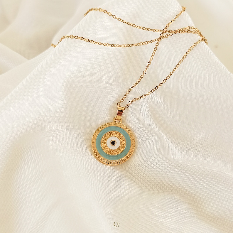 Puede incluir: Un collar de cadena dorada con un colgante redondo que presenta un ojo azul maligno con un centro blanco y detalles dorados.