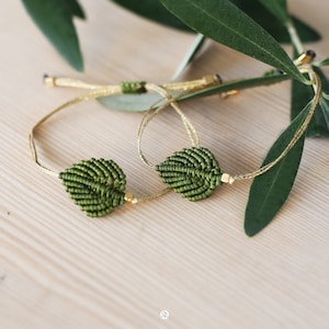 Può includere: Due braccialetti dell'amicizia a forma di foglia verde con perline dorate. I braccialetti sono realizzati con corda annodata e sono legati insieme.