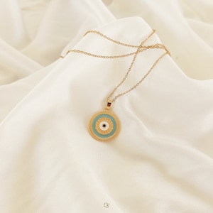 Puede incluir: Collar de cadena dorada con un colgante redondo con un dise&ntilde;o de ojo azul malvado. El colgante tiene un centro blanco y detalles dorados.
