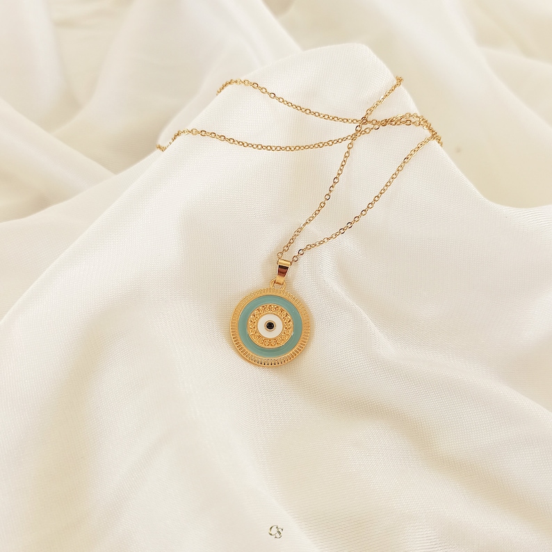 Puede incluir: Collar de cadena dorada con un colgante redondo con un dise&ntilde;o de ojo azul malvado. El colgante tiene un centro blanco y detalles dorados.