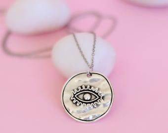 Puede incluir: Collar de plata con un colgante redondo con un diseño de ojo turco. El colgante tiene una superficie texturizada y una pequeña perla en el centro del ojo.