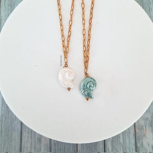 May include: Two seashell pendant necklaces. One pendant is white, the other is teal. Both pendants are attached to gold-colored chain necklaces. The necklaces are displayed on a white, round surface.