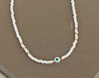 Puede incluir: Collar de cuentas blancas con un dije de ojo turco dorado. El collar está sobre una superficie marrón.