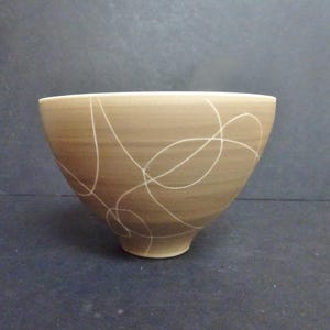 May include: A ceramic bowl with a light brown exterior and a white interior rim. The bowl features a series of white, free-form lines that swirl around the exterior. The bowl has a rounded shape with a slightly flared rim.
