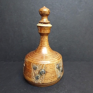 May include: A brown ceramic bottle with a decorative stopper. The bottle has a round base with textured horizontal lines and square and circular designs. The stopper is a lighter brown colour and has a rounded top.