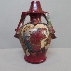 May include: A red ceramic vase with a detailed design featuring figures and floral elements. The vase has a unique shape with handles and a flared top. The artwork includes shades of blue, beige, and green, adding to its artistic appeal.