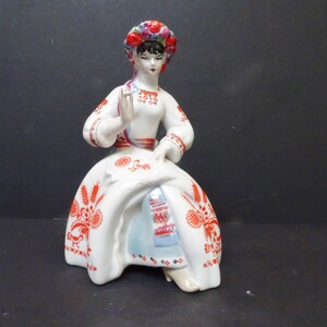 May include: A ceramic figurine of a woman in traditional dress. The figure wears a white dress with red floral patterns and a floral headpiece. The figurine is posed in a seated position.