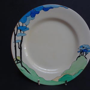 May include: A round ceramic plate with a cream-coloured centre and a hand-painted landscape design along the rim. The design features black-outlined green hills, a light tan path, and stylised blue trees with dark blue and light blue foliage.