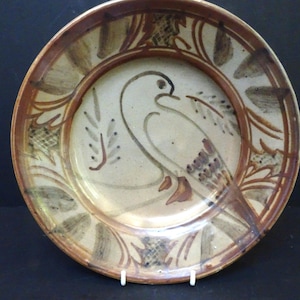 May include: A round ceramic plate featuring a hand-painted brown bird illustration in the center. The rim is decorated with a repeating pattern of brown leaves and cross-hatched details against a cream-colored background with a glossy finish.