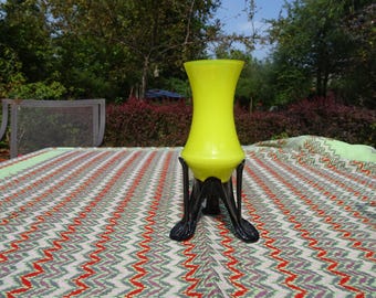 May include: A bright yellow glass vase with a flared top and a black, ornate metal stand. The vase is set on a patterned tablecloth with red, green, and white zigzags. The background includes trees and foliage.