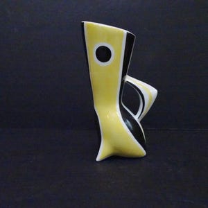 Puede incluir: Un jarrón de cerámica con una silueta alta y angular en colores amarillo y negro. El diseño incluye un prominente acento circular negro en la sección superior amarilla y un asa curva que se extiende desde el lateral del cuerpo.