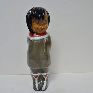 Könnte beinhalten: Eine Keramikfigur einer Person mit schwarzen Haaren und einem braunen Gesicht. Die Figur trägt einen braunen Mantel mit weißem und rotem Kragen und eine weiße Hose mit roten Akzenten. Die Figur steht aufrecht.