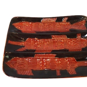 Puede incluir: Una fuente rectangular de cerámica negra con tres diseños de peces en relieve de color rojo anaranjado. Los peces tienen escamas y aletas detalladas, y la fuente tiene un acabado brillante. Adecuada para servir aperitivos o platos pequeños.