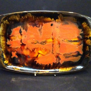 May include: An oval ceramic serving platter featuring a glossy glaze with a vibrant orange centre that transitions into deep brown and golden yellow tones along the edges. The surface displays a mottled, reactive finish with dark, flowing patterns.