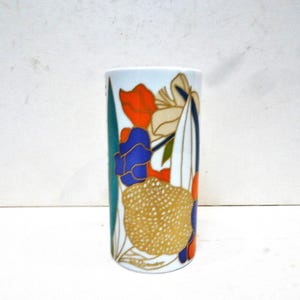 Puede incluir: Un jarrón cilíndrico con un diseño floral. El jarrón presenta un fondo blanco con flores coloridas en naranja, azul y dorado. El jarrón tiene una franja turquesa en un lado.