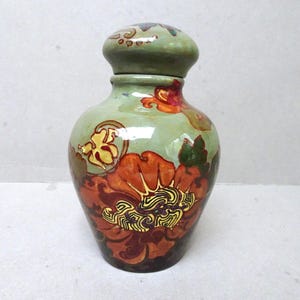 May include: A decorative ceramic vase with a lid. The vase is painted with a floral design in shades of orange, yellow, and green. The vase is approximately 8 inches tall.