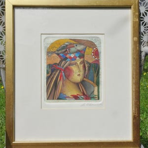 May include: A framed art print featuring a stylized portrait of a person with a fish headdress. The artwork has a warm color palette with reds, yellows, and blues. The print is in a gold-colored frame.